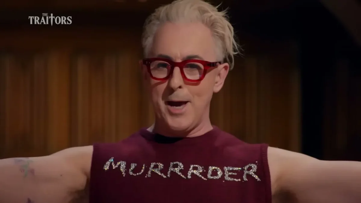 Alan Cumming, host of The Traitors, in red glasses and a sleeveless top that reads MURRRDER