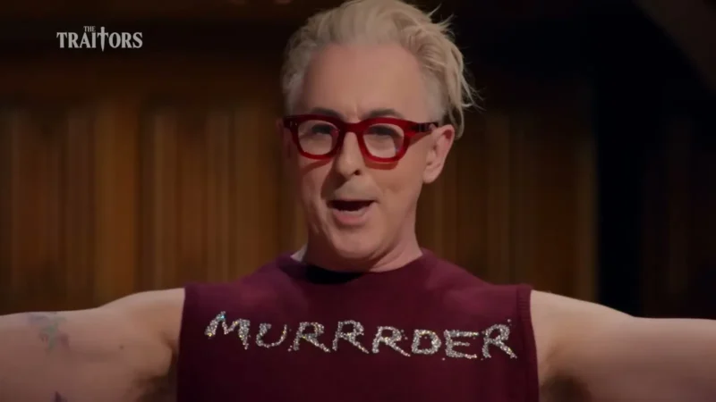 Alan Cumming, host of The Traitors, in red glasses and a sleeveless top that reads MURRRDER