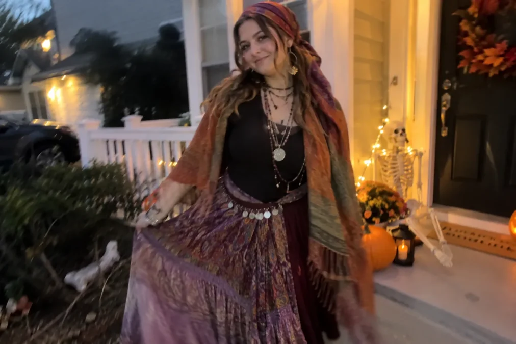 Illustration of a woman dressed as a fortune teller in a headscarf, layered necklaces, a fringed shawl and a long paisley skirt on a porch decorated for Halloween