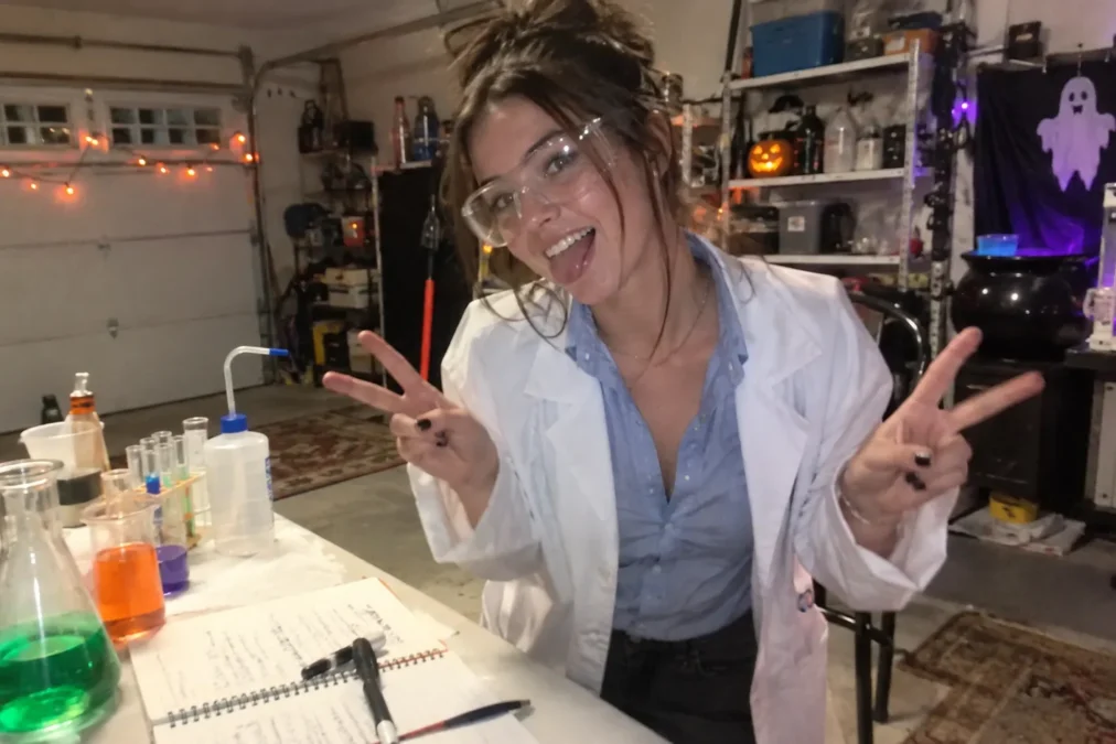 Illustration of a woman dressed as a scientist in a white lab coat, blue button-down and safety glasses, behind a table of colorful flasks