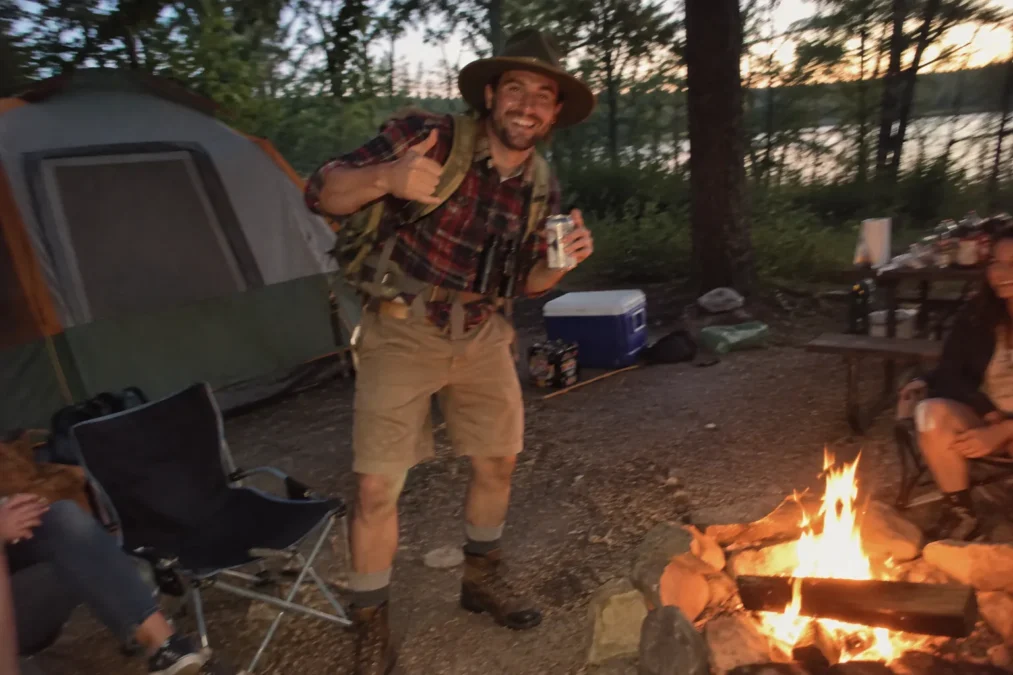 Illustration of a man dressed as a camper in a wide-brim hat, red plaid flannel, khaki shorts, hiking boots and a backpack by a campfire