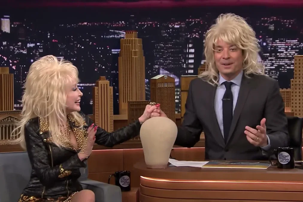 Jimmy Fallon wearing a curly blonde wig next to Dolly Parton and an empty wig stand on The Tonight Show