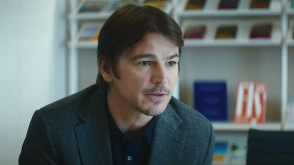 Josh Hartnett in a gray blazer talking across a desk in an office lined with books