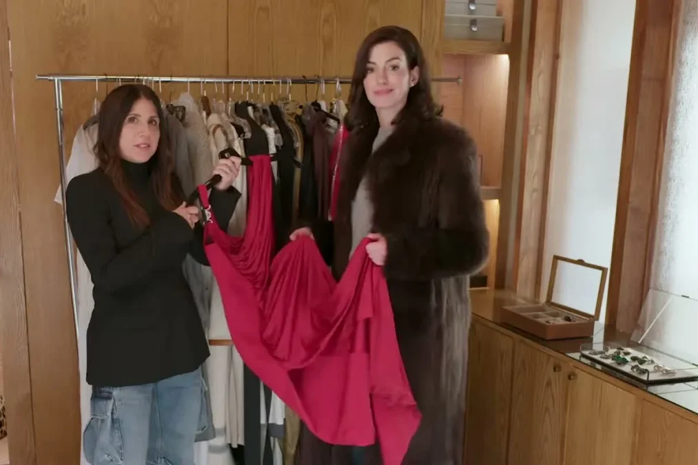 Anne Hathaway in a fur coat holding up a red dress beside costume designer Jacqueline Demeterio in a walk-in closet