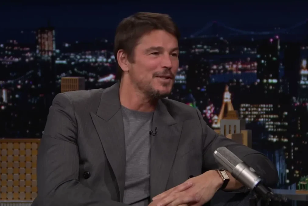 Josh Hartnett in a gray suit smiling on the Tonight Show couch with the New York skyline behind him