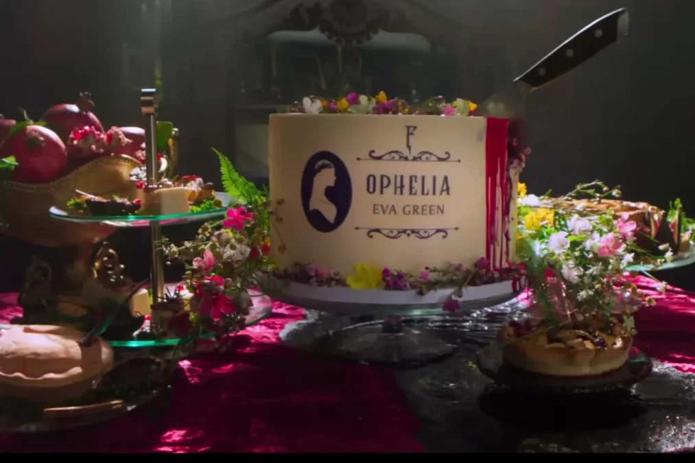 A cake with a place card reading Ophelia, Eva Green, on a banquet table with flowers, fruit and a knife stuck in the cake
