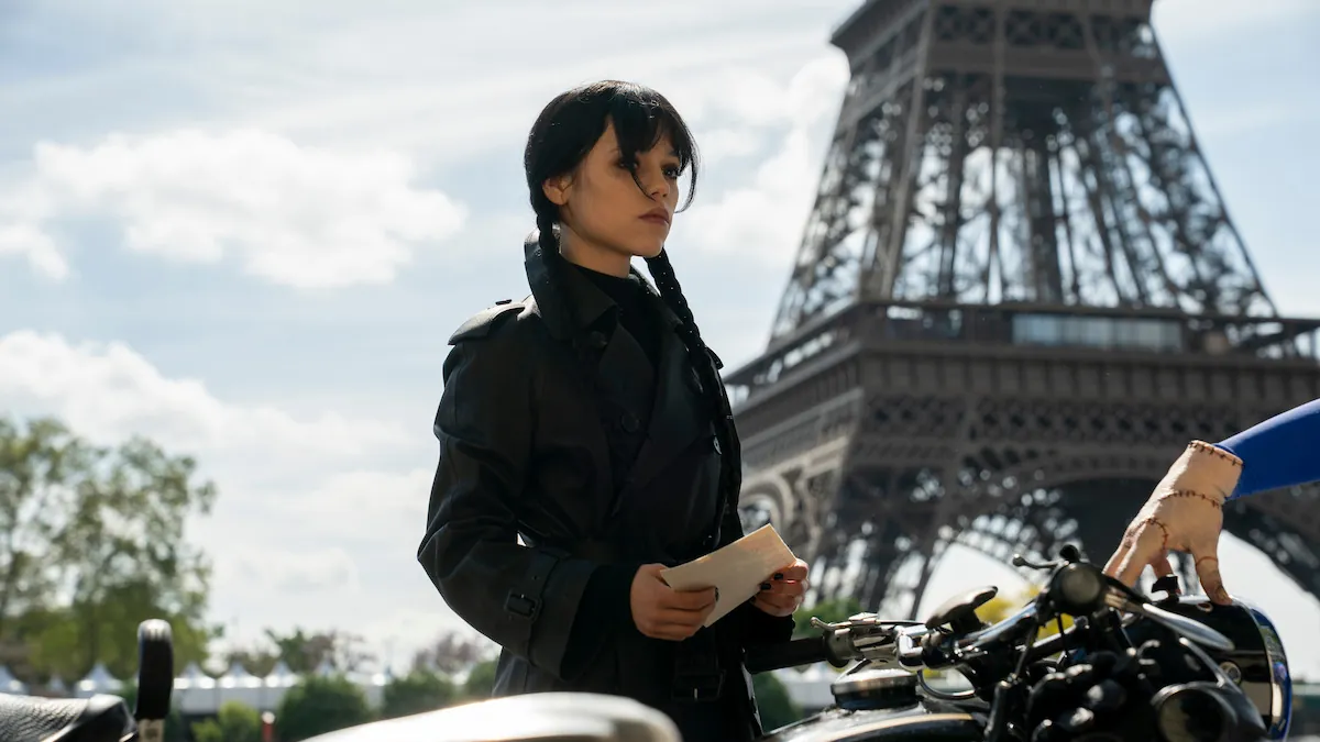 Jenna Ortega as Wednesday Addams in a black coat, holding papers beside a row of motorcycles with the Eiffel Tower behind her