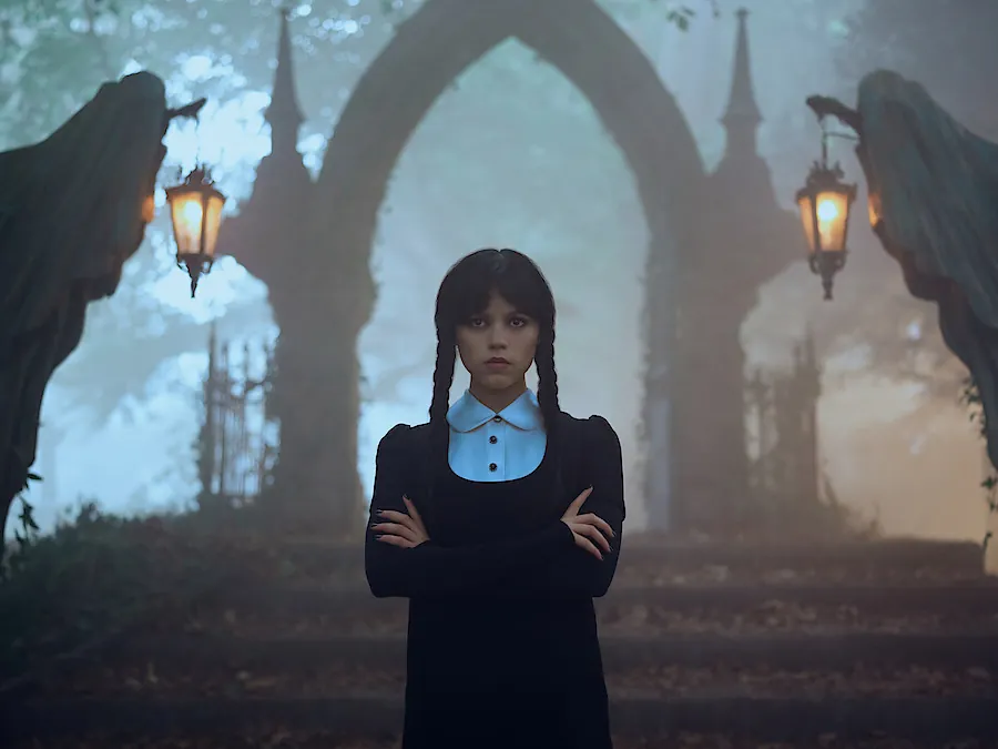Jenna Ortega as Wednesday Addams with crossed arms in a misty graveyard under a stone arch