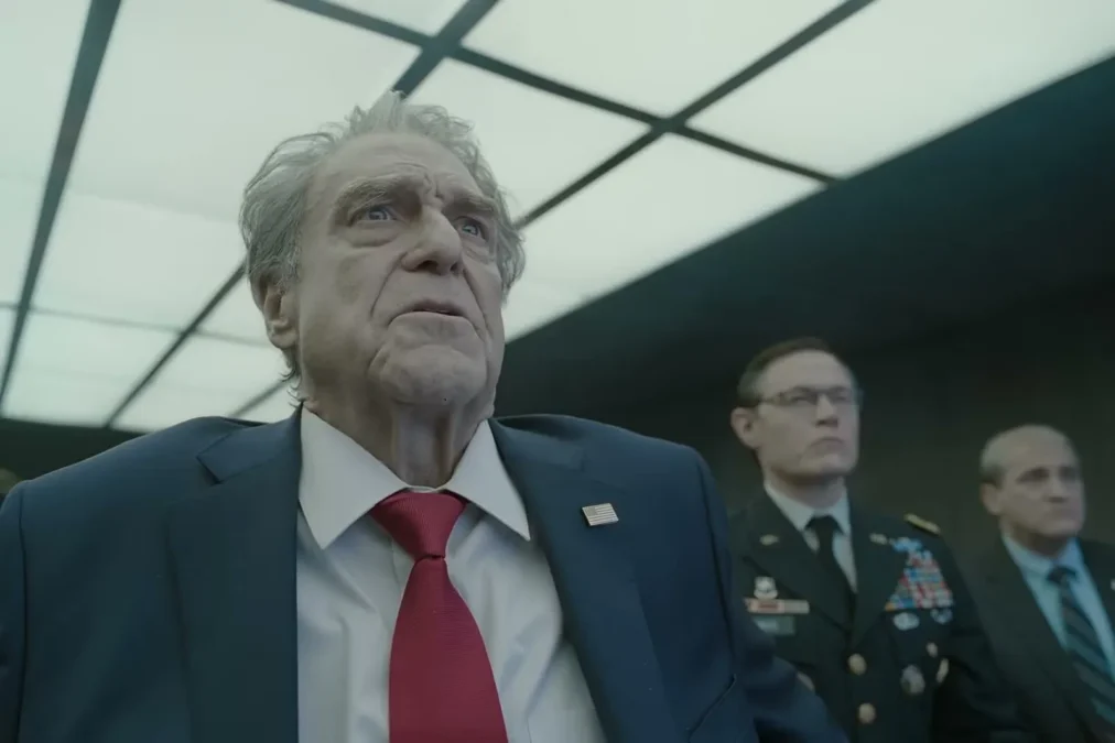 A gray-haired man in a navy suit and red tie stands with military officers in a war room in the Digger trailer