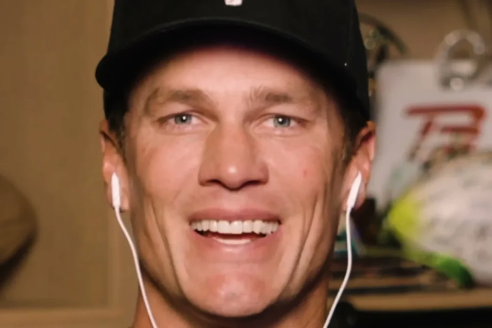 Tom Brady smiles in a black cap and earbuds while answering fan questions in a FOX Sports video