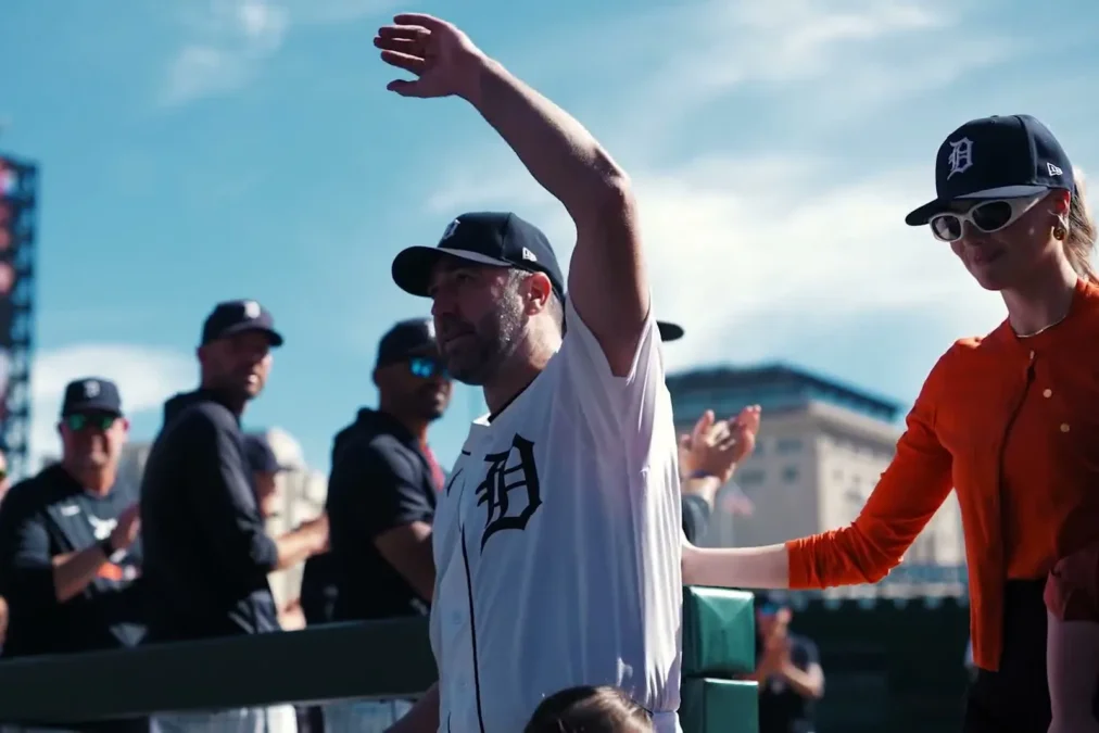 Justin Verlander in his Tigers jersey waves to the crowd at Comerica Park as Kate Upton, in an orange cardigan, Tigers cap and white sunglasses, stands beside him with a hand on his back