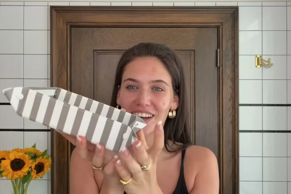 Sienna Spiro smiling without makeup in a tiled bathroom, holding a striped makeup bag, in a Vogue beauty video