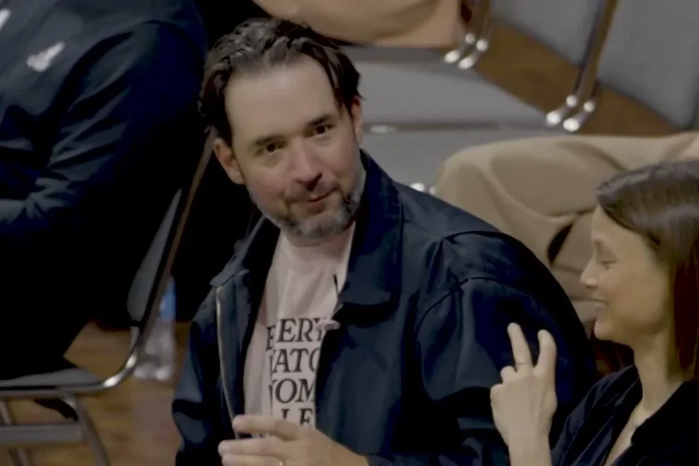Alexis Ohanian in a dark jacket sitting courtside at a LOVB volleyball match, next to two-time Olympian Courtney Thompson