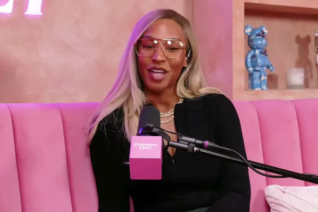 Savannah James in glasses and a black top at a microphone on the pink set of her podcast Everybody's Crazy