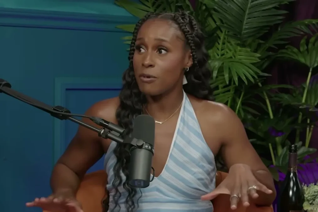 Issa Rae in a striped halter top talking at a microphone on a podcast set with plants and a teal wall