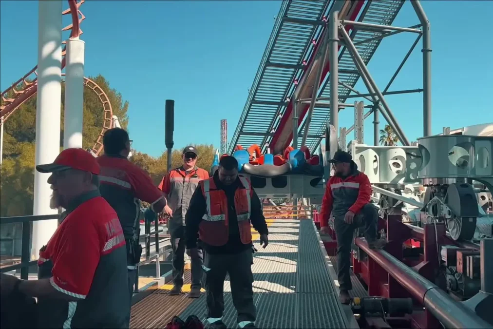 Six Flags Magic Mountain crew members in red work shirts on the X2 station platform, with the ride's lift structure behind them