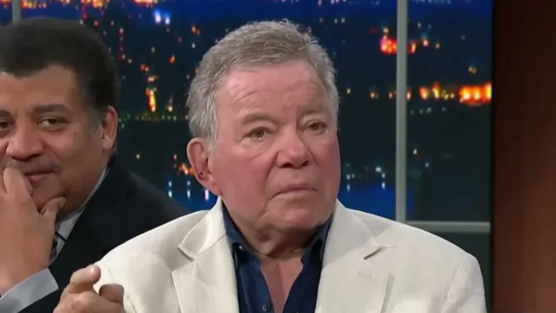 William Shatner in a white linen jacket and dark shirt on a late-night talk show set