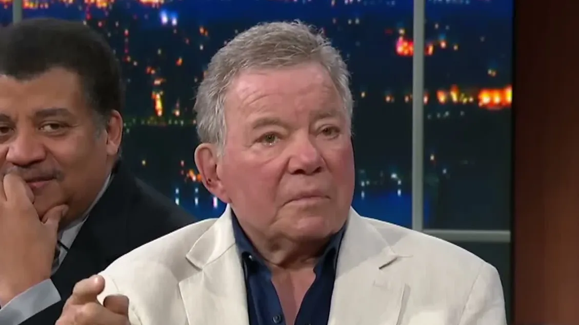 William Shatner in a white linen jacket and dark shirt on a late-night talk show set