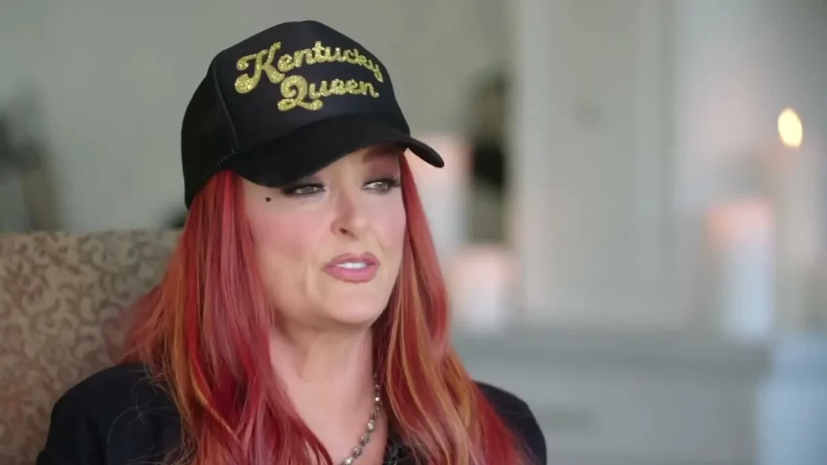 Wynonna Judd in a black Kentucky Queen cap talking in a living room