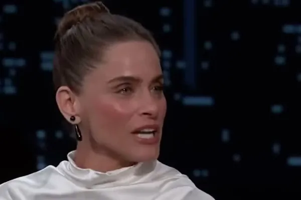 Amanda Peet with her hair in a bun and a white satin top talking on Jimmy Kimmel Live