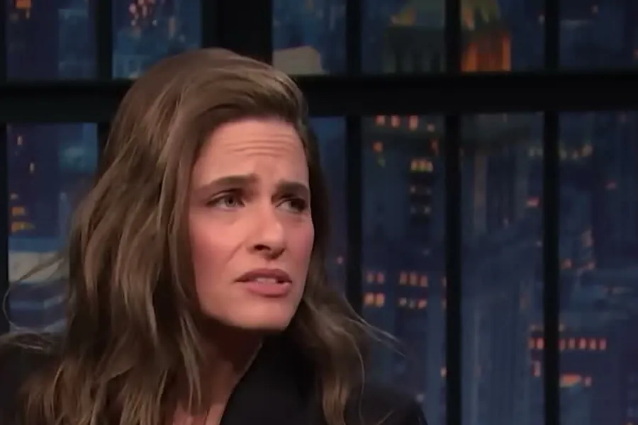 Amanda Peet with long brown hair in a black blazer talking on the Late Night with Seth Meyers set