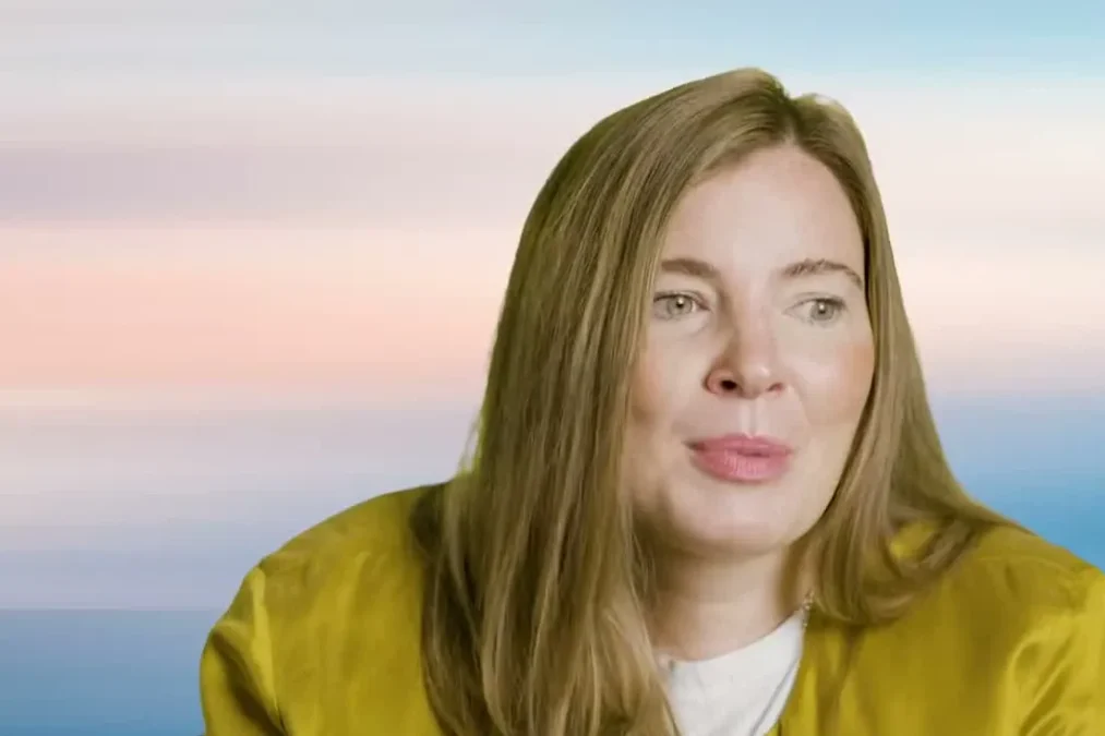 Brooke Eby in a mustard-colored jacket in front of an ocean-colored backdrop in the Muscular Dystrophy Association's tribute video