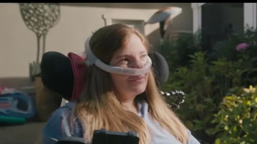 Brooke Eby smiling in a wheelchair outdoors with a nasal breathing mask in the Salesforce video