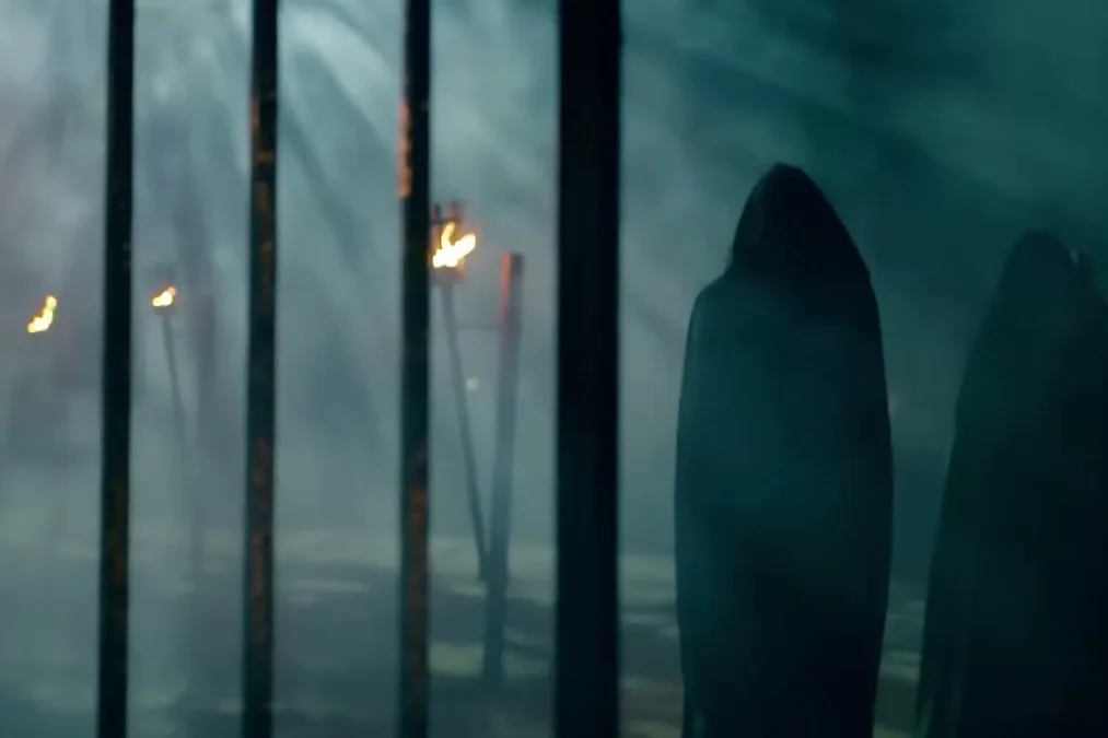 Two figures in dark hooded cloaks stand behind iron bars in a smoky courtyard lit by torches