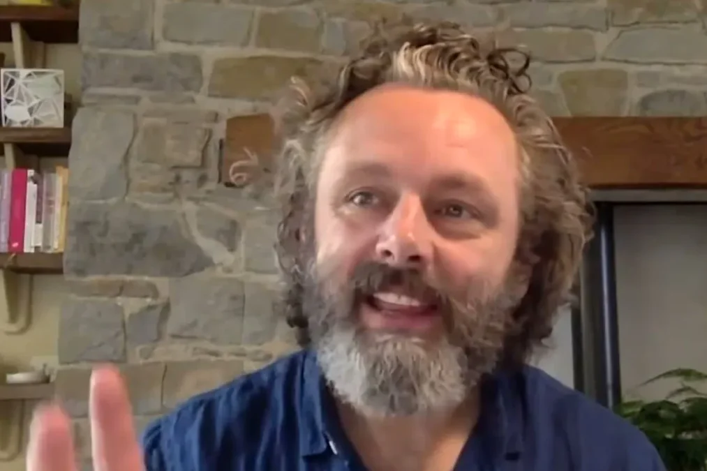 Michael Sheen with curly hair and a gray beard in a blue shirt talking on a video call in front of a stone wall