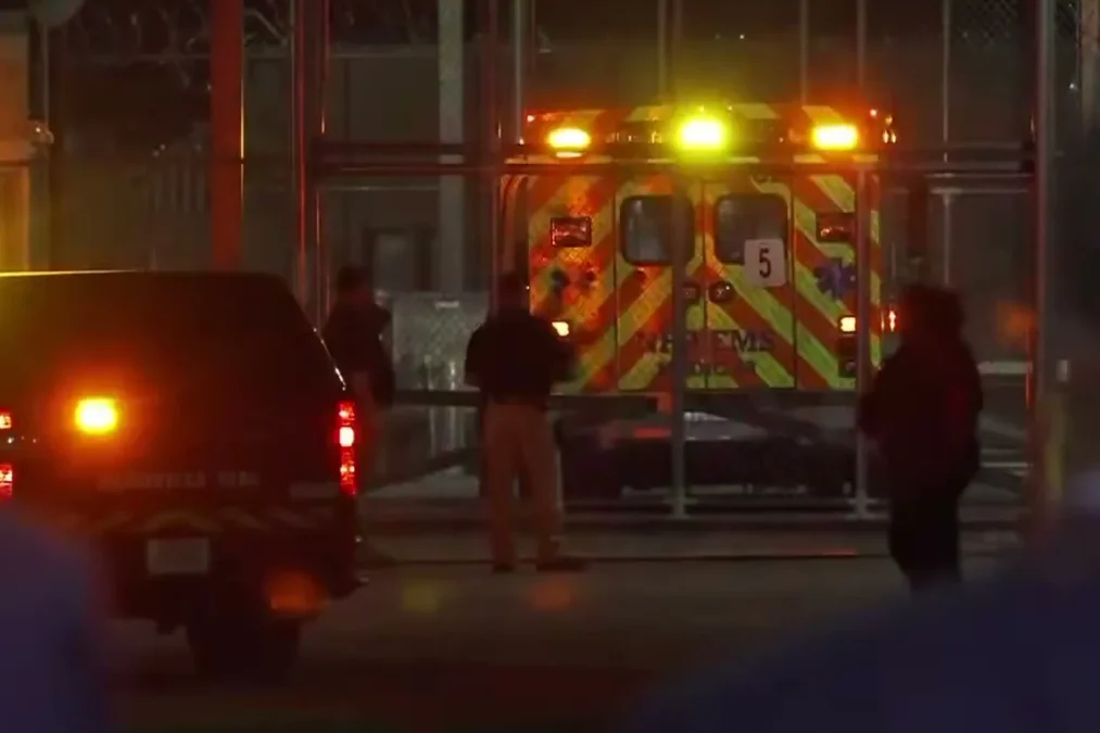 An ambulance with flashing lights behind a prison security fence at night, with corrections staff standing nearby