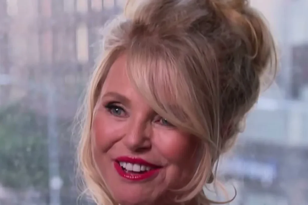 Christie Brinkley with blond hair in an updo and a black strap top smiling in front of a city window