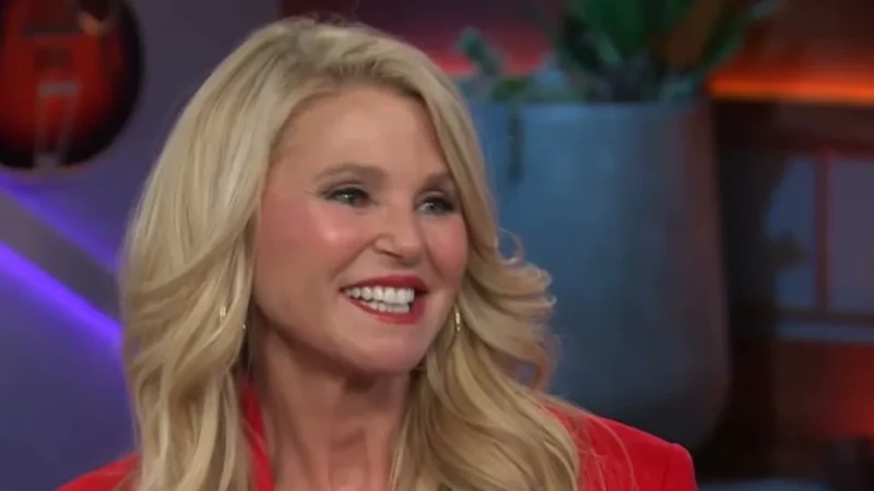 Christie Brinkley with long wavy blond hair and a red blazer smiling on the set of The Kelly Clarkson Show