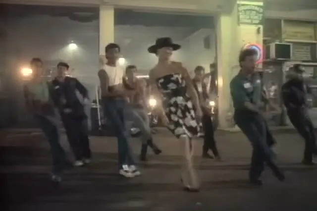 Dancers in work shirts and a woman in a black-and-white dress and black hat at a gas station at night in the Uptown Girl music video
