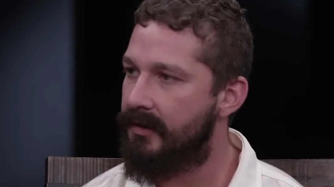 Shia LaBeouf with curly dark hair and a beard in a white shirt looking to the side during an interview