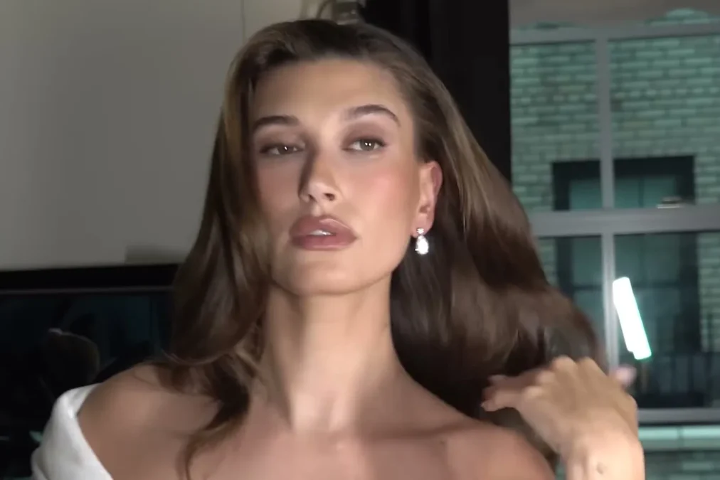 Hailey Bieber with long brown hair, pearl earrings and a white robe having her hair styled before the Met Gala