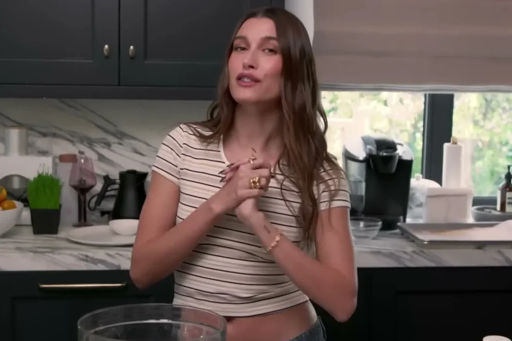 Hailey Bieber in a striped T-shirt standing in her kitchen gesturing while she talks