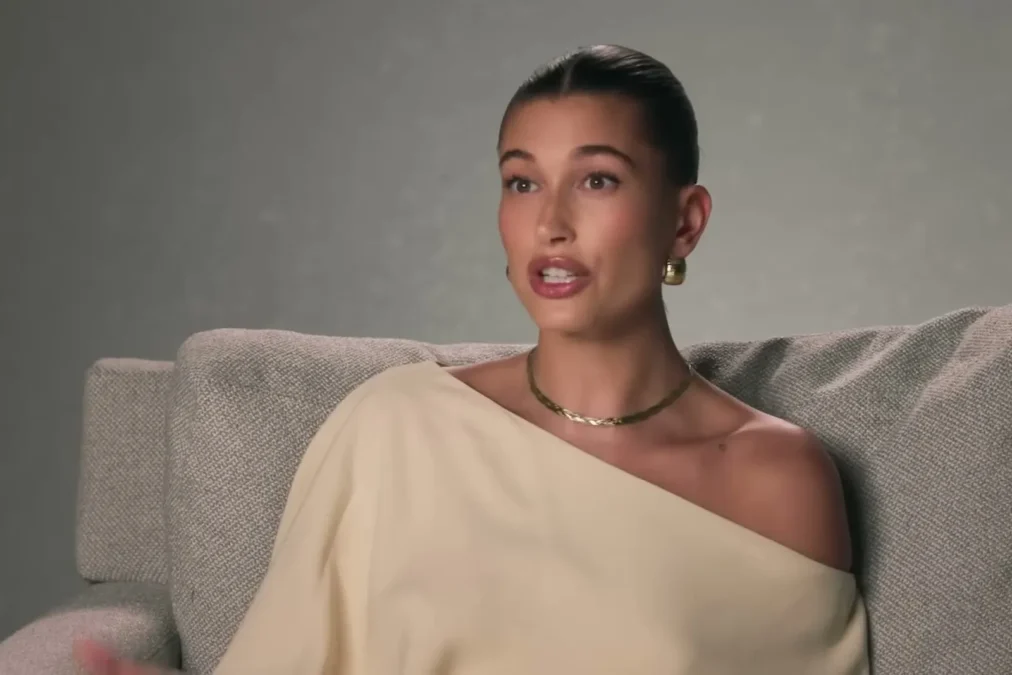 Hailey Bieber in a cream off-the-shoulder sweater and a gold necklace speaking while seated on a gray couch