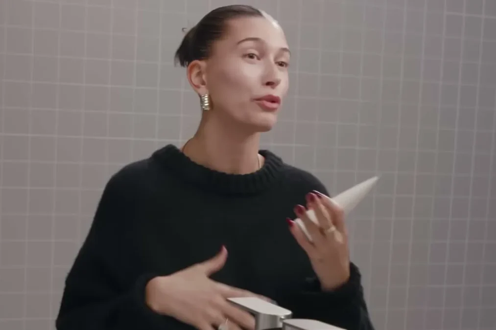 Hailey Bieber in a black sweater with her hair in a bun talking in front of a round mirror in a gray-tiled bathroom