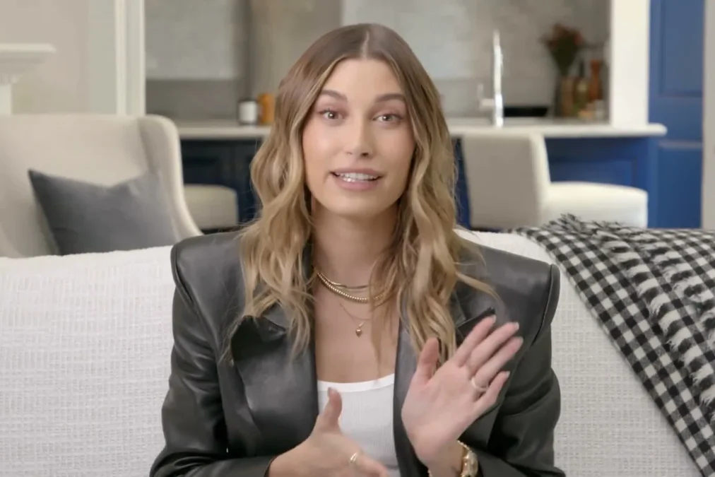 Hailey Bieber with wavy blond-brown hair in a black leather blazer smiling on a white couch