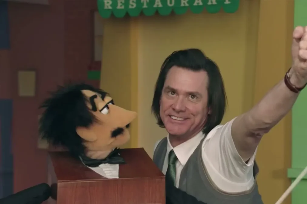 Jim Carrey in a vest and tie, smiling, raising a fist next to a puppet on a children's-show set in Kidding