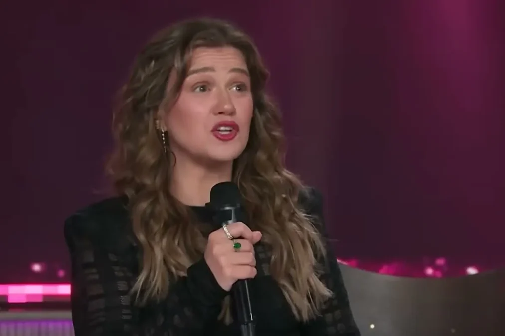 Kelly Clarkson with long wavy brown hair holding a microphone and speaking on the stage of The Kelly Clarkson Show
