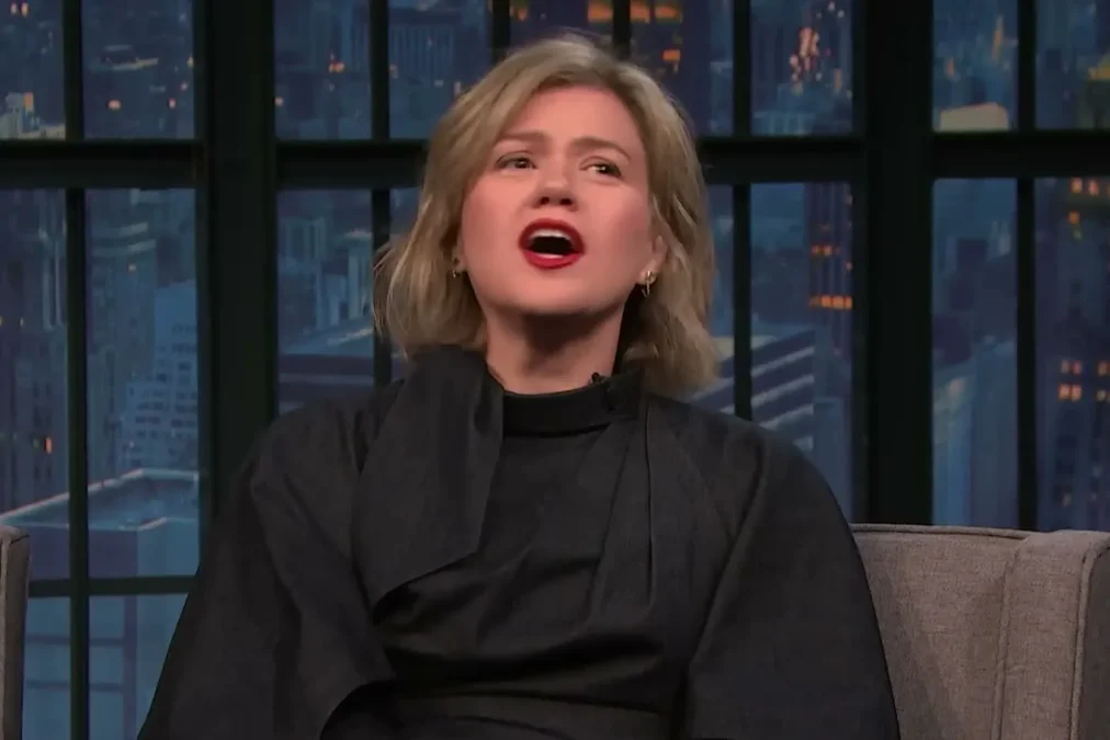 Kelly Clarkson with a short blond bob and red lipstick in a black top talking on the Late Night with Seth Meyers set
