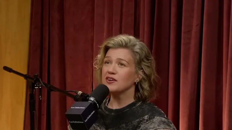 Kelly Clarkson with short wavy hair sitting on an orange couch in front of a red velvet curtain, talking at the Las Culturistas podcast microphone