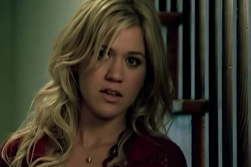 Kelly Clarkson with long blond wavy hair in a red cardigan looking at the camera in the Because of You video