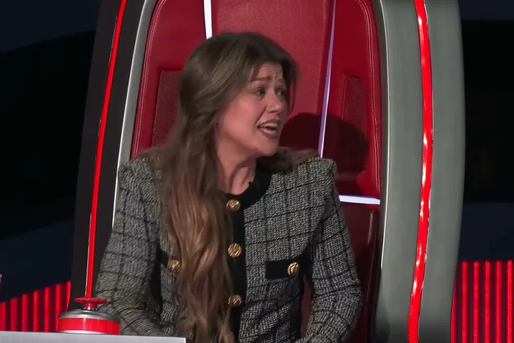 Kelly Clarkson in a gray tweed jacket sitting in a red coach's chair on The Voice