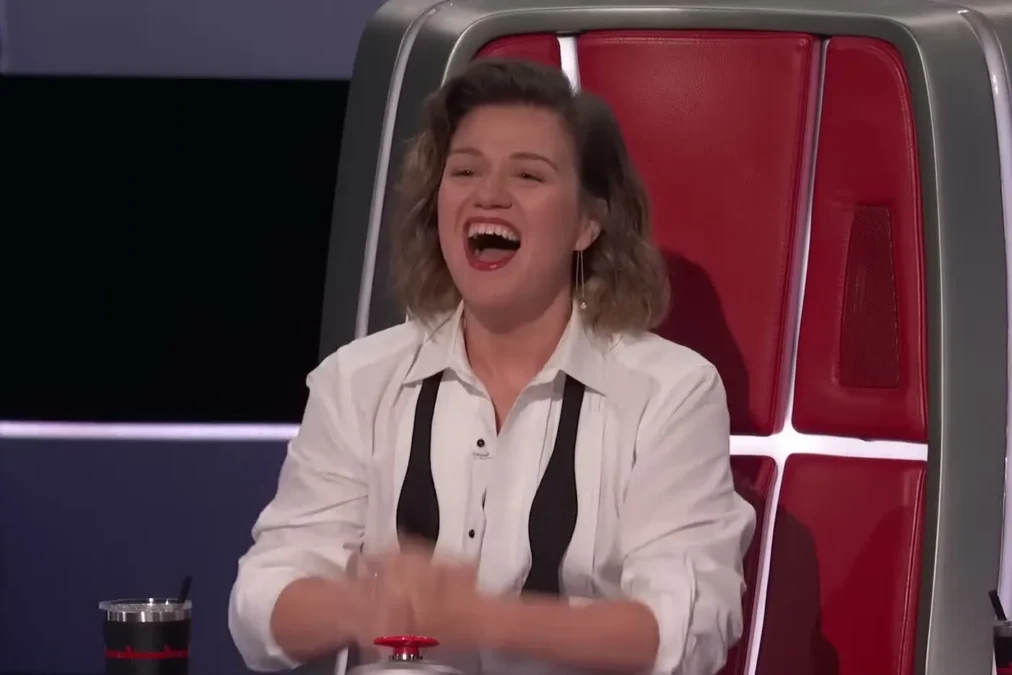 Kelly Clarkson in a white shirt and black suspenders laughing hard in her red chair with her hands on the buzzer on The Voice