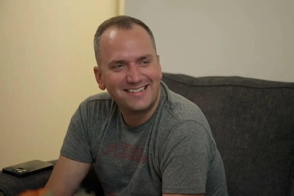 Ken Urker laughing in a gray T-shirt on a couch in a clip from Gypsy Rose: Life After Lock Up