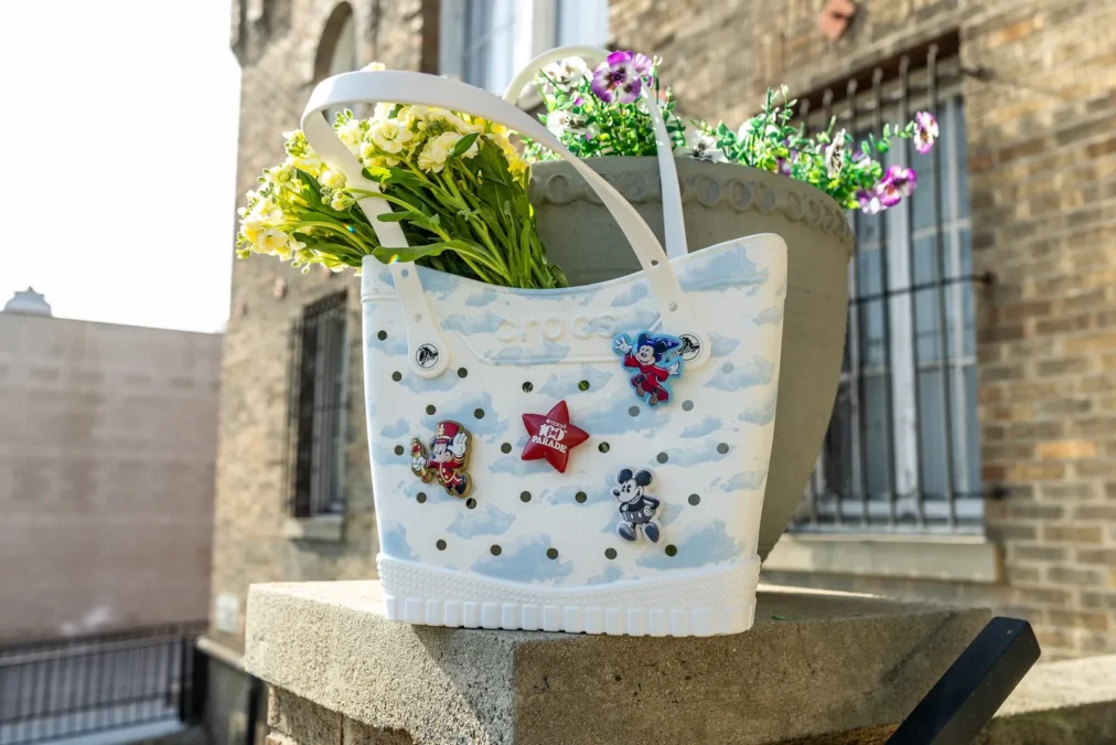 A white Crocs tote bag printed with blue clouds and decorated with Mickey and star Jibbitz charms, holding flowers