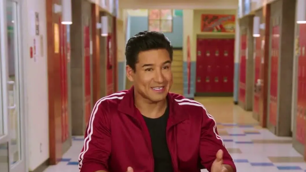 Mario Lopez in a maroon track jacket sitting in a high school hallway during an interview