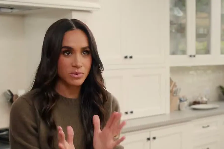 Meghan Markle with long wavy dark hair in an olive sweater gesturing with both hands in a white kitchen
